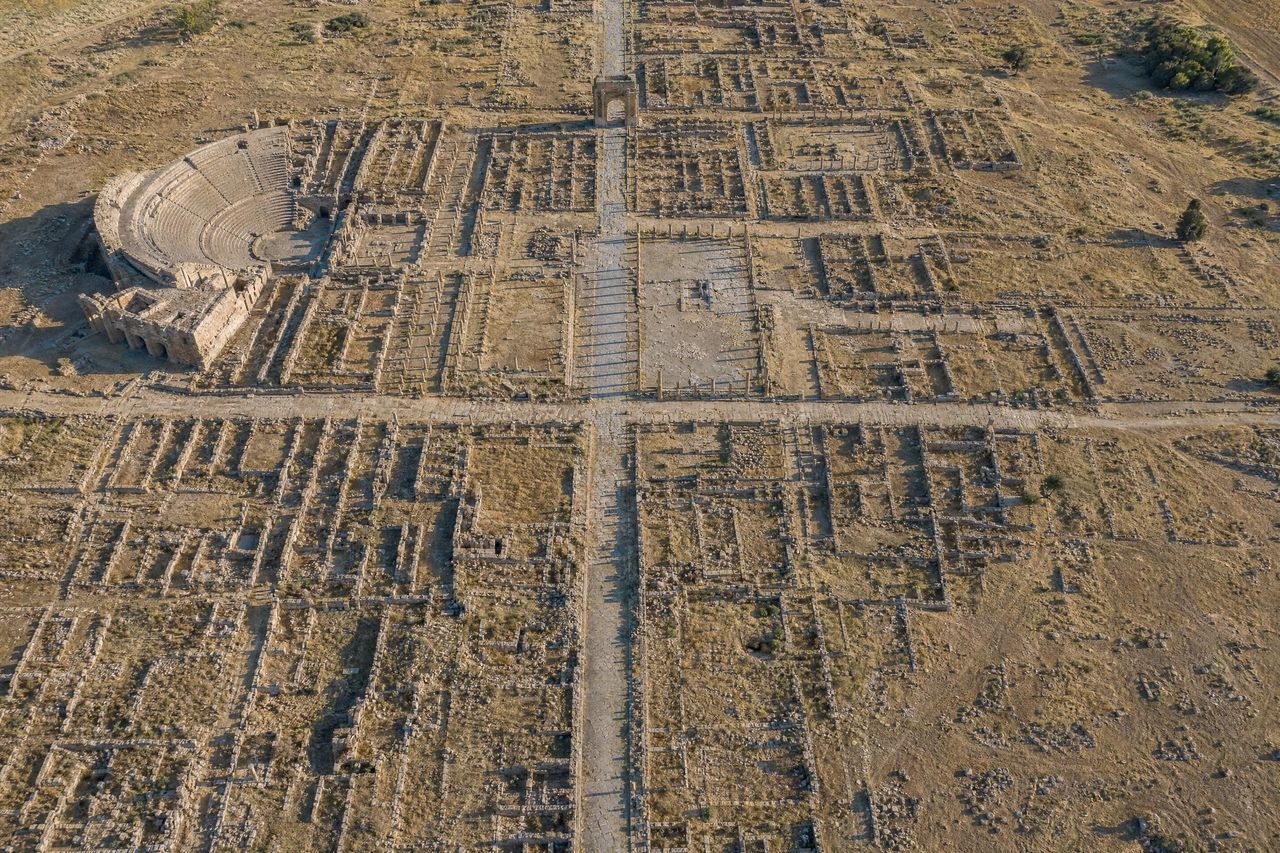 Vista aérea de las ruinas de una ciudad romana trazada en cuadrícula, con el teatro, el foro y un arco al final de la calle principal (imagen generada con AI)