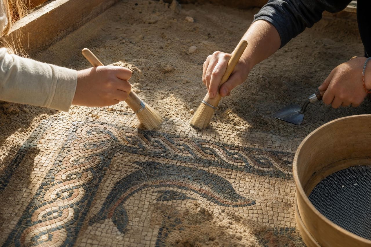 Manos de dos niños limpiando con pinceles un mosaico de estilo romano en un taller de arqueología (imagen generada con AI)